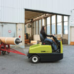 Operator driving a Clark CTX electric tow tractor outside an industrial facility while towing a cart carrying a large cylindrical roll, demonstrating material transport between production and loading areas.