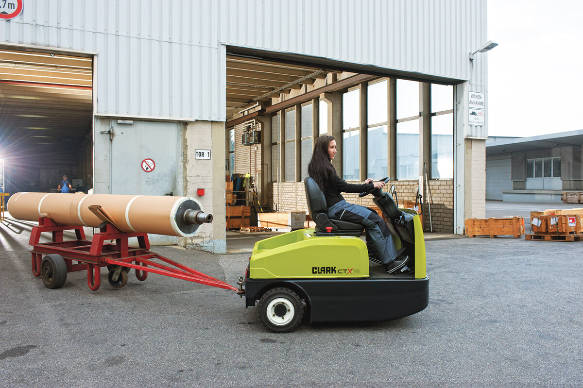 Operator driving a Clark CTX electric tow tractor outside an industrial facility while towing a cart carrying a large cylindrical roll, demonstrating material transport between production and loading areas.