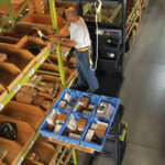 Warehouse employee using a Clark OSX order picker to select inventory from storage bins, with organized parts trays on a pallet platform and high-density warehouse shelving in the background.