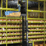 Worker operating a Clark OSX order picker at an elevated height in a warehouse, accessing inventory from high storage racks while standing on a raised operator platform with a pallet of boxed products.
