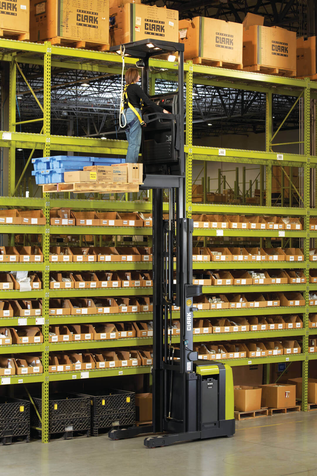 Worker operating a Clark OSX order picker at an elevated height in a warehouse, accessing inventory from high storage racks while standing on a raised operator platform with a pallet of boxed products.