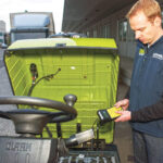 Technician inspecting and charging a Clark CTX electric tow tractor, with the battery compartment open and charging equipment connected in an outdoor industrial loading area.
