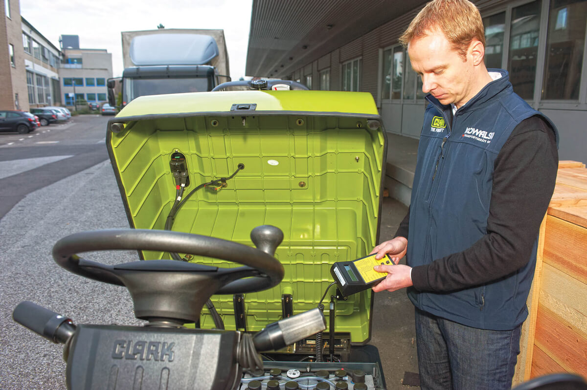 Technician inspecting and charging a Clark CTX electric tow tractor, with the battery compartment open and charging equipment connected in an outdoor industrial loading area.
