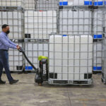 Worker using a Clark electric pallet jack to transport a large liquid storage container (IBC tote) through a warehouse, surrounded by stacked industrial containers and storage inventory.
