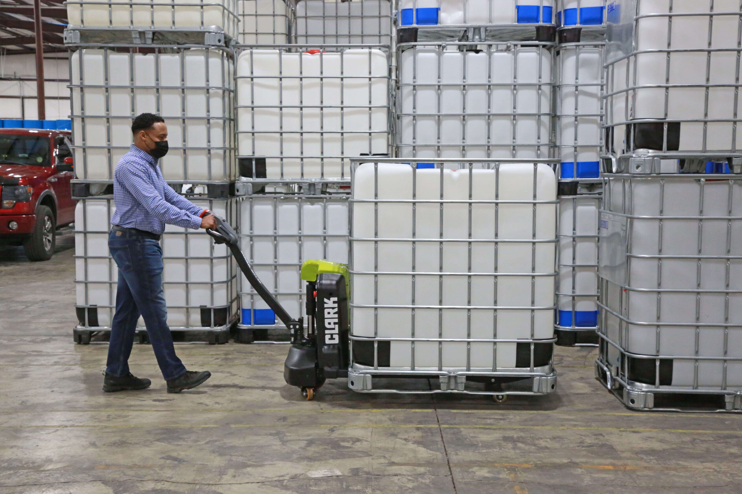 Worker using a Clark electric pallet jack to transport a large liquid storage container (IBC tote) through a warehouse, surrounded by stacked industrial containers and storage inventory.