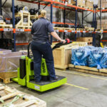 Worker operating a Clark ride-on electric pallet truck in a warehouse, transporting palletized materials through a storage area with industrial components, crates, and shelving.