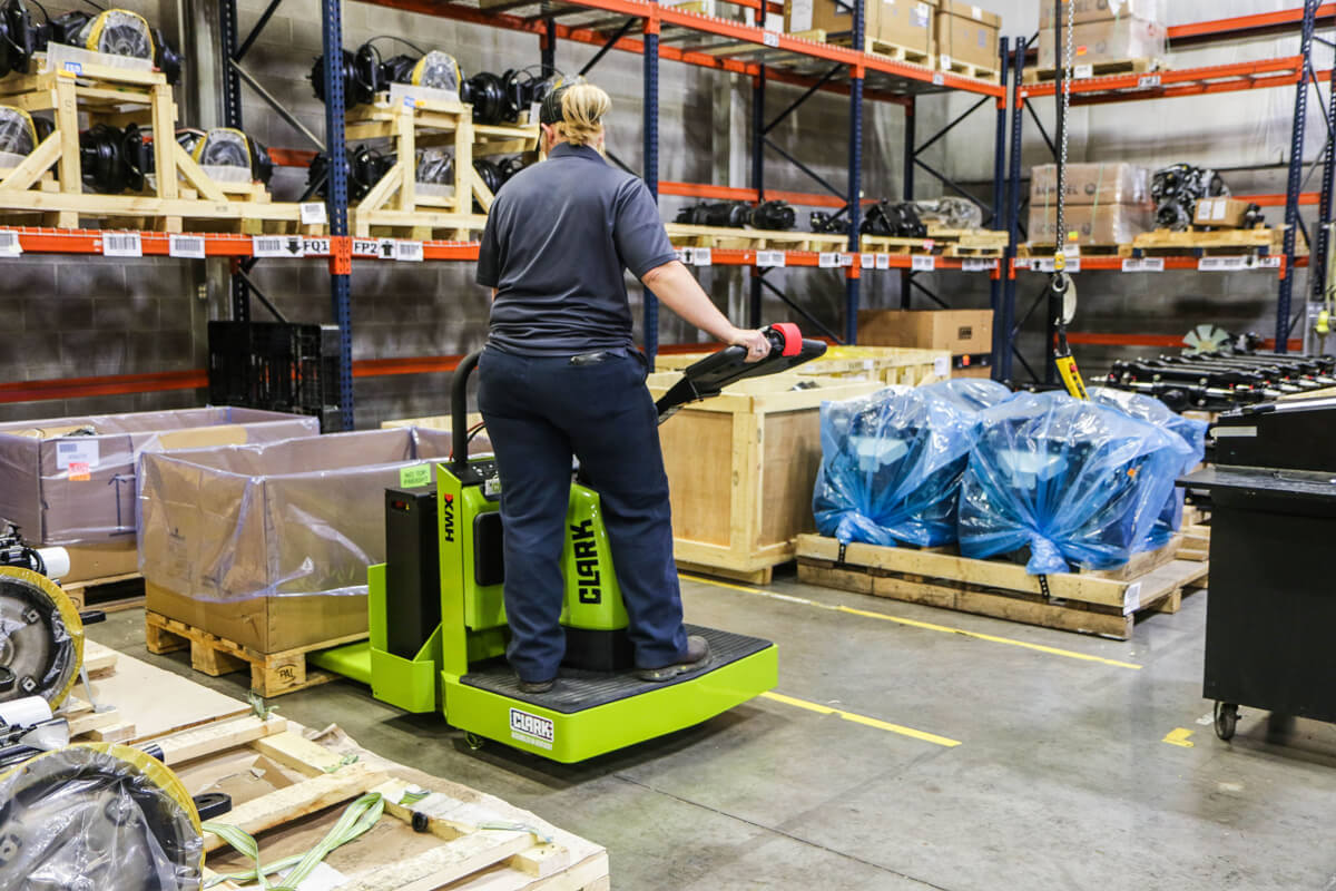 Worker operating a Clark ride-on electric pallet truck in a warehouse, transporting palletized materials through a storage area with industrial components, crates, and shelving.