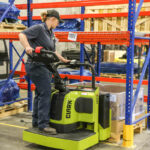 Warehouse worker operating a Clark ride-on electric pallet truck while positioning a palletized container in a storage area, surrounded by industrial racking and inventory.