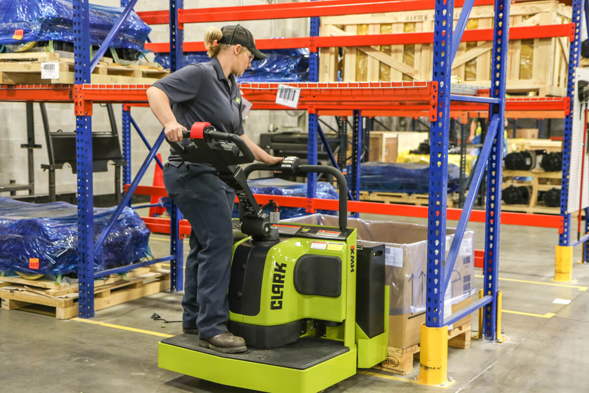 Warehouse worker operating a Clark ride-on electric pallet truck while positioning a palletized container in a storage area, surrounded by industrial racking and inventory.
