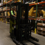 Warehouse worker operating a Clark electric reach truck in a storage aisle, surrounded by industrial shelving stocked with parts, equipment, and palletized inventory.