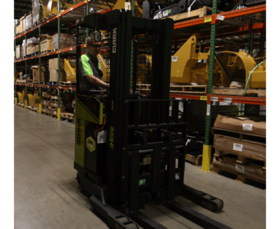Warehouse worker operating a Clark electric reach truck in a storage aisle, surrounded by industrial shelving stocked with parts, equipment, and palletized inventory.