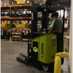 Operator driving a Clark NPX 17 order picker through a warehouse, with industrial racking, palletized inventory, and equipment stored throughout the facility.