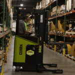 Warehouse employee operating a Clark NPX 17 order picker between storage racks, retrieving inventory from shelving in a large parts distribution warehouse.