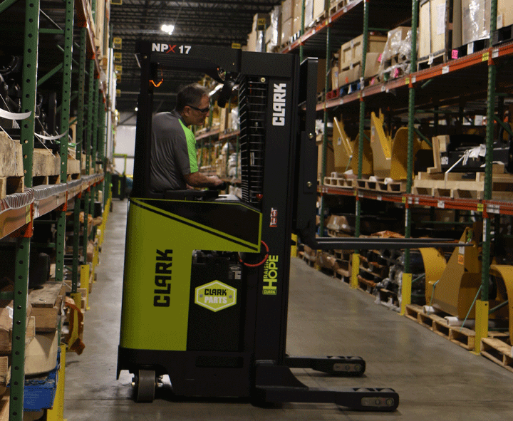 Warehouse employee operating a Clark NPX 17 order picker between storage racks, retrieving inventory from shelving in a large parts distribution warehouse.