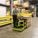 Worker operating a Clark ride-on electric pallet truck while transporting a loaded pallet through a warehouse aisle, with safety railings, storage racks, and industrial inventory in the background.