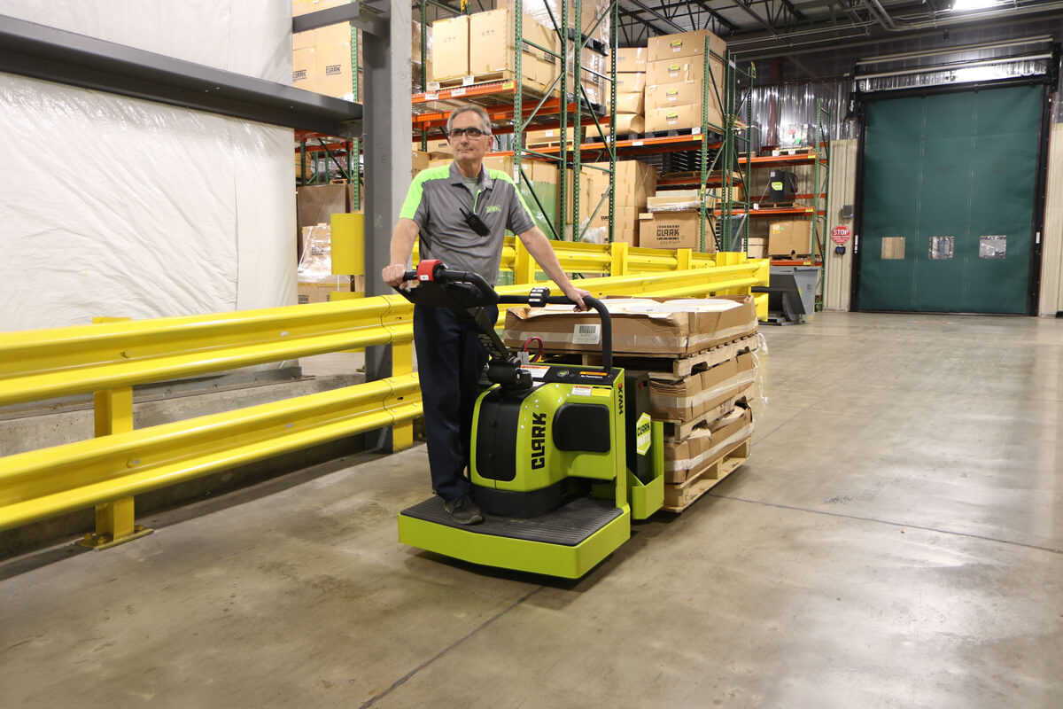 Worker operating a Clark ride-on electric pallet truck while transporting a loaded pallet through a warehouse aisle, with safety railings, storage racks, and industrial inventory in the background.