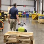 Worker operating a Clark WPL40 electric pallet jack while transporting stacked wooden pallets through a warehouse, viewed from behind on an open industrial floor.