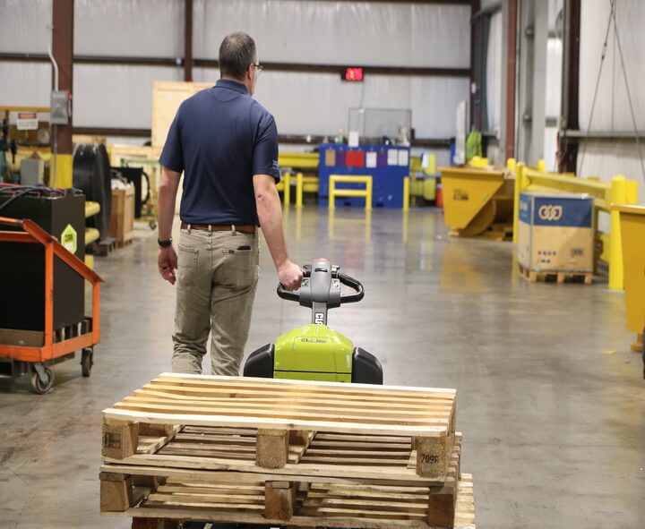 Worker operating a Clark WPL40 electric pallet jack while transporting stacked wooden pallets through a warehouse, viewed from behind on an open industrial floor.