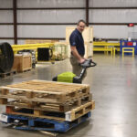 Worker maneuvering a Clark WPL40 electric pallet jack while transporting stacked wooden pallets across a warehouse floor, with industrial equipment and storage areas visible in the background.
