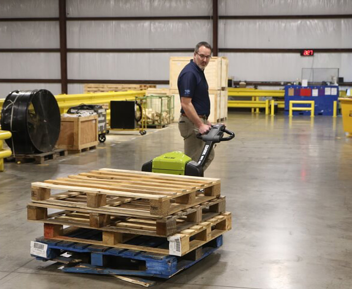 Worker maneuvering a Clark WPL40 electric pallet jack while transporting stacked wooden pallets across a warehouse floor, with industrial equipment and storage areas visible in the background.