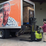 Worker operating a Clark electric walkie stacker to load palletized boxes into the back of a delivery truck at a warehouse loading dock.