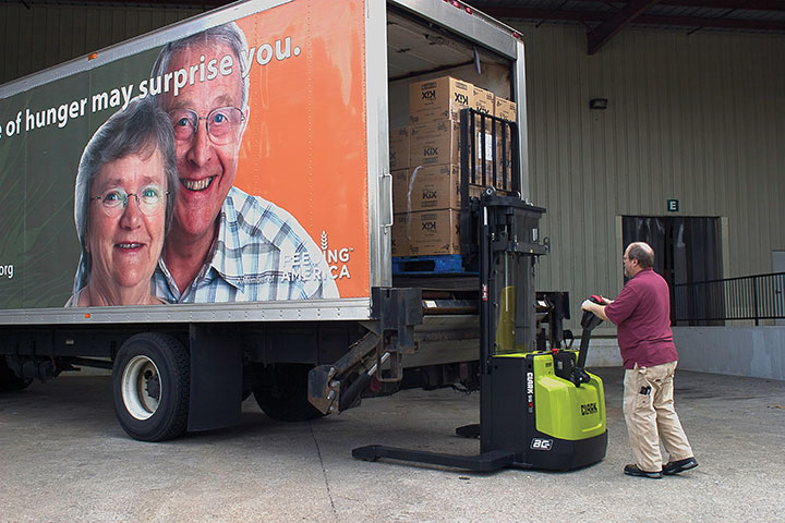 Worker operating a Clark electric walkie stacker to load palletized boxes into the back of a delivery truck at a warehouse loading dock.