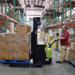 Warehouse worker operating a Clark SSX electric walkie stacker to transport and position a pallet of boxed inventory in a narrow warehouse aisle lined with tall storage racks and palletized goods.