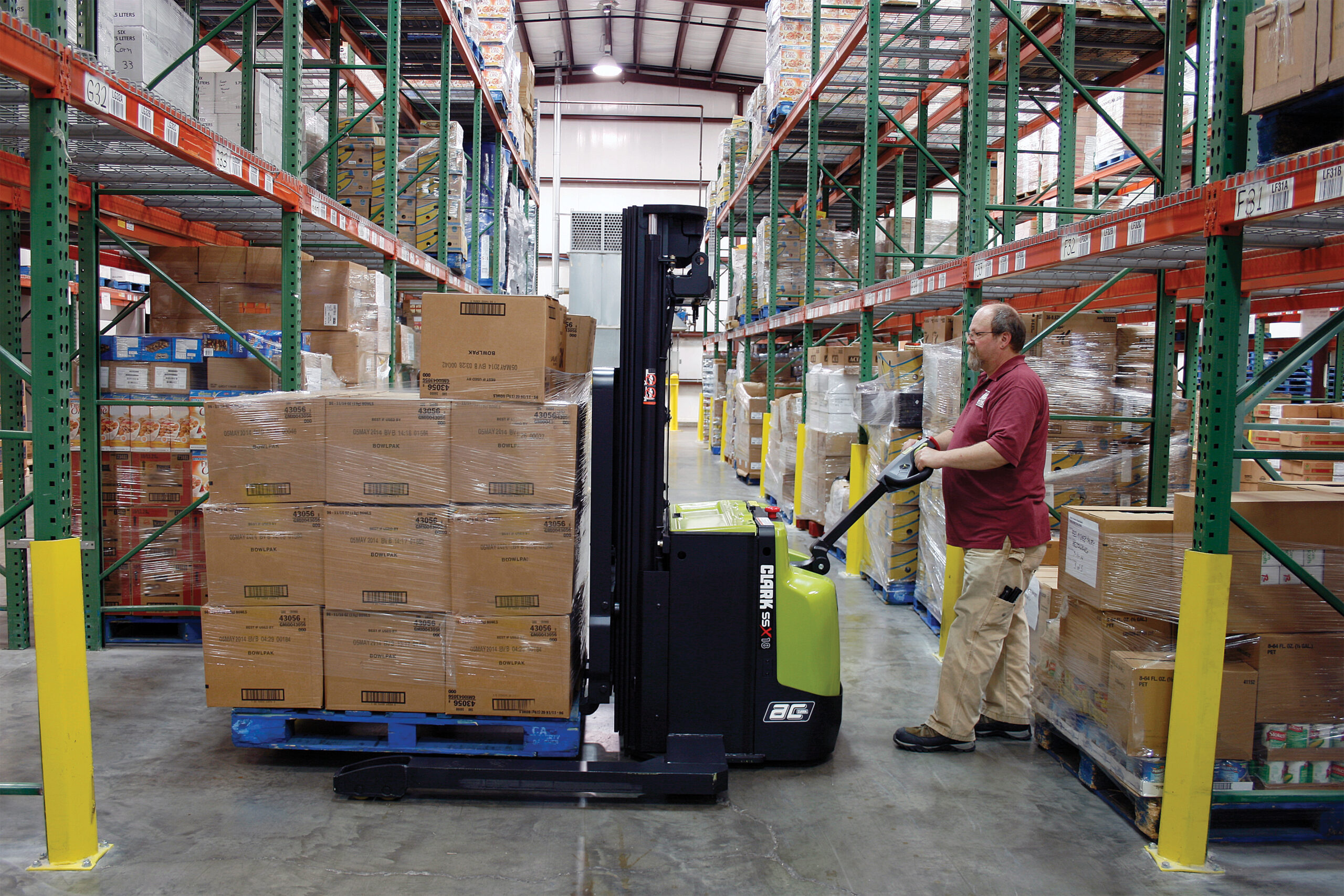 Warehouse worker operating a Clark SSX electric walkie stacker to transport and position a pallet of boxed inventory in a narrow warehouse aisle lined with tall storage racks and palletized goods.
