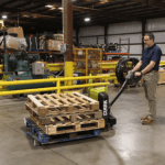 Worker using a Clark WPL40 electric pallet jack to transport stacked wooden pallets across an industrial warehouse floor, with machinery, storage racks, and safety barriers in the background.