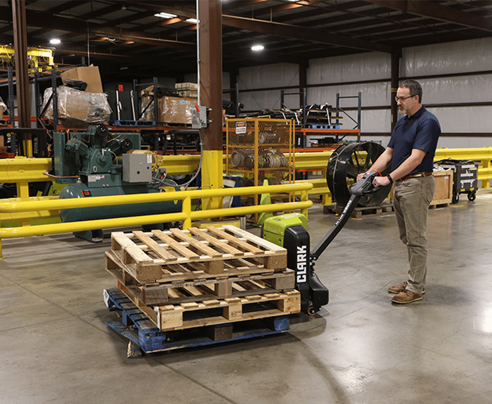 Worker using a Clark WPL40 electric pallet jack to transport stacked wooden pallets across an industrial warehouse floor, with machinery, storage racks, and safety barriers in the background.